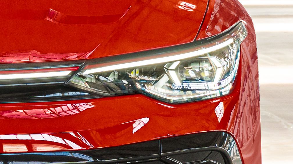 Detailný záber červeného Golfu GTI so zameraním na jeden svetlomet. Povrch je lesklý a odráža svetlo. Svetlomet má moderný dizajn s ostrými hranami.