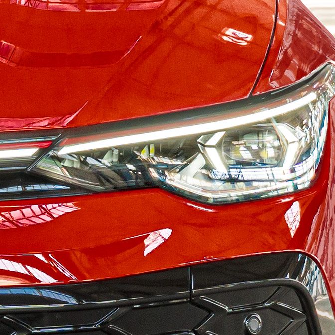 Detailný záber červeného Golfu GTI so zameraním na jeden svetlomet. Povrch je lesklý a odráža svetlo. Svetlomet má moderný dizajn s ostrými hranami.