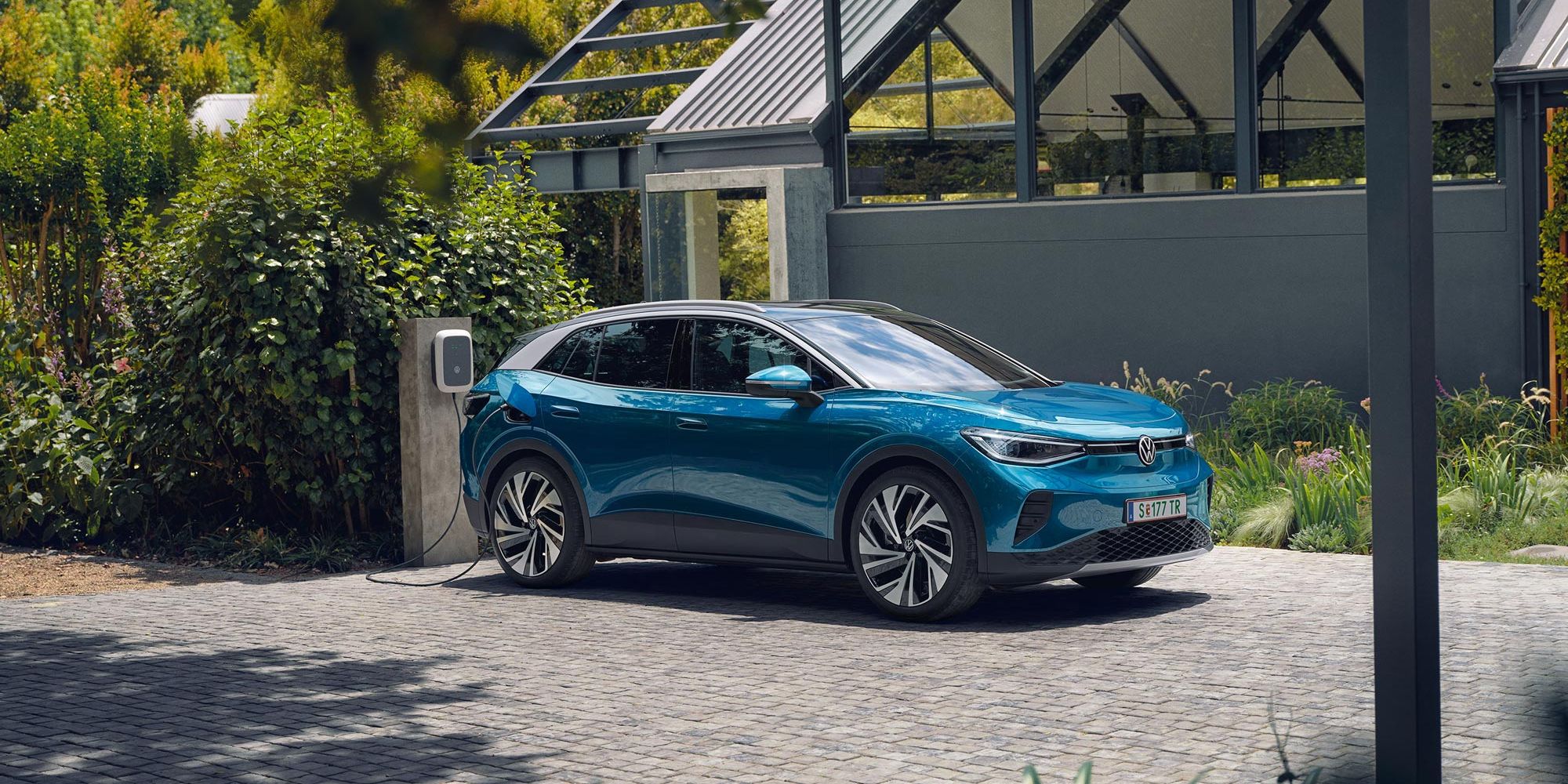 Modré elektrické SUV značky Volkswagen stojí na dláždenej ploche pri domácej nabíjacej stanici. V pozadí je zeleň a malá moderná budova.