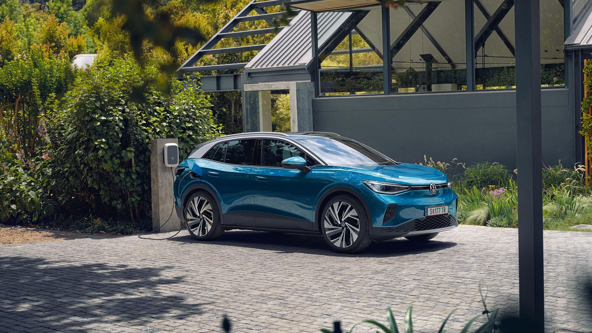 Modré elektrické SUV značky Volkswagen stojí na dláždenej ploche pri domácej nabíjacej stanici. V pozadí je zeleň a malá moderná budova.