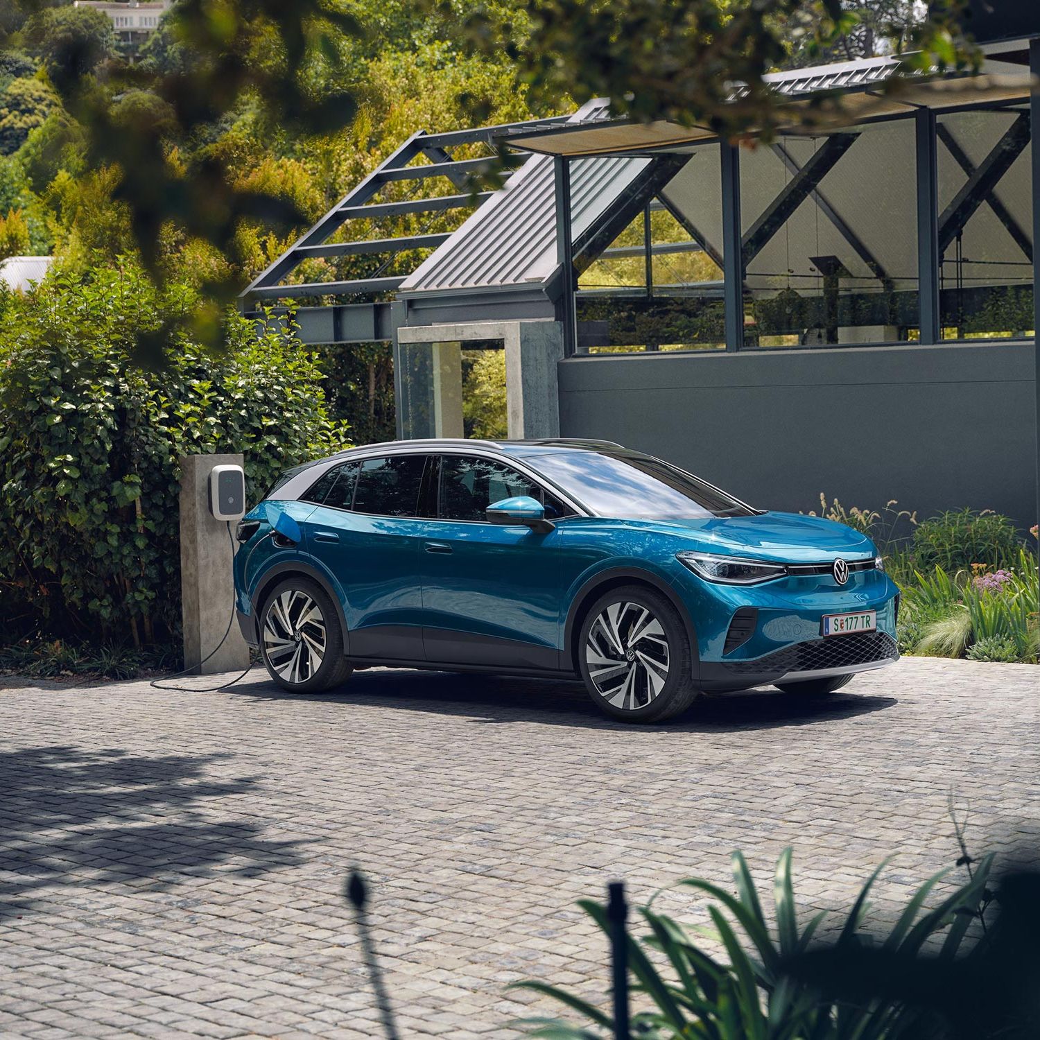 Modré elektrické SUV značky Volkswagen stojí na dláždenej ploche pri domácej nabíjacej stanici. V pozadí je zeleň a malá moderná budova.