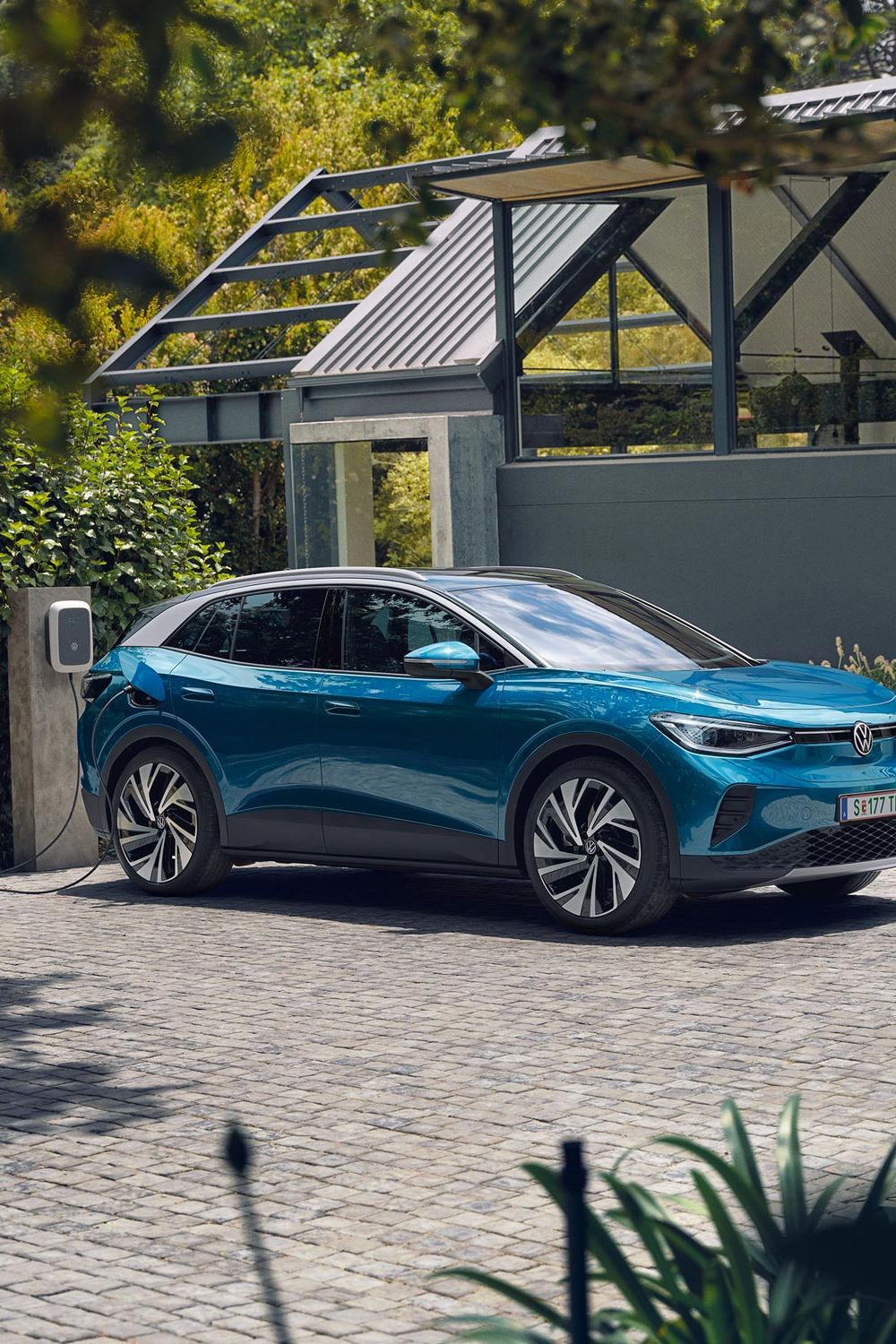Modré elektrické SUV značky Volkswagen stojí na dláždenej ploche pri domácej nabíjacej stanici. V pozadí je zeleň a malá moderná budova.