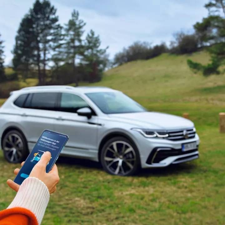 Žena sa pozerá na svoj smartfón. V pozadí stojí jej vozidlo Volkswagen na zelenom parkovisku uprostred vidieckej krajiny.