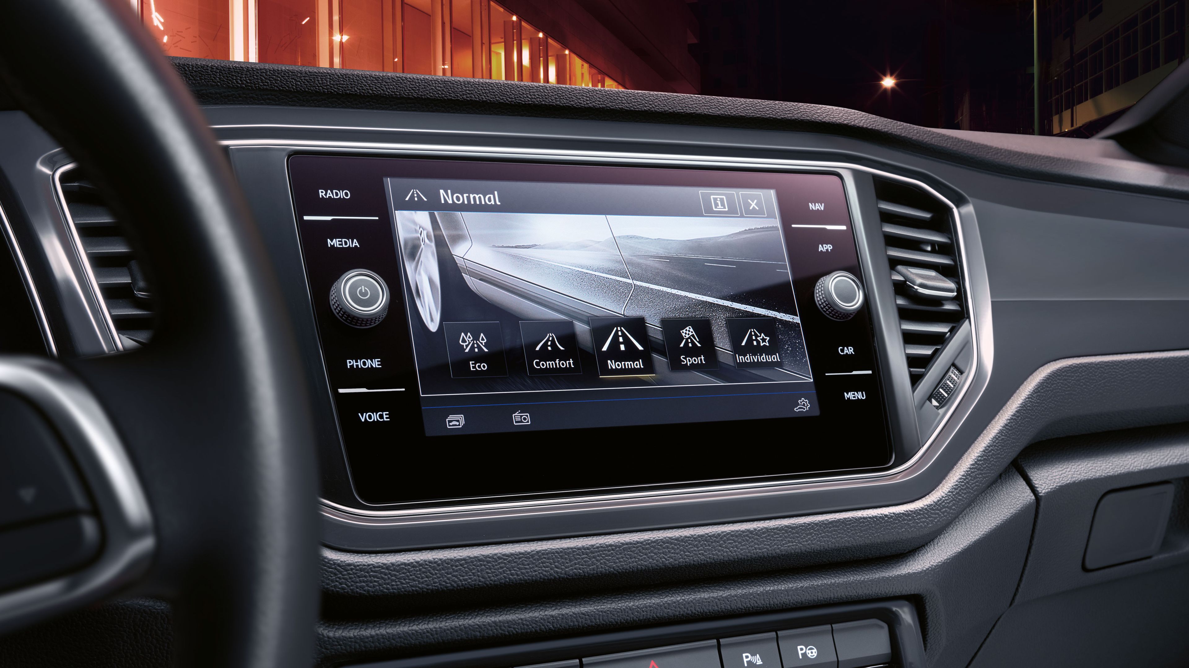 Dotykový displej systému infotainment zobrazuje rôzne jazdné režimy, ako je ekologický, komfortný, normálny a športový. Vľavo sa nachádza volant. Okolo obrazovky sú tlačidlá ovládania rádia, médií a iných funkcií.
