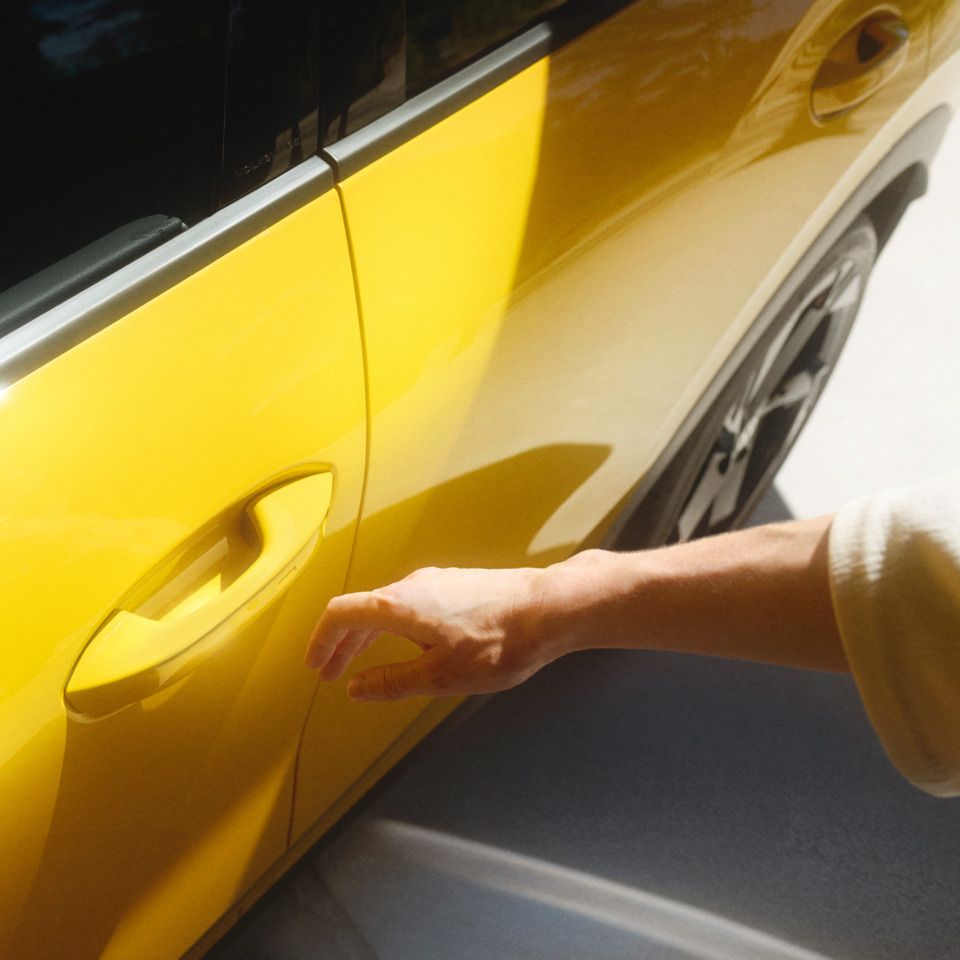 Človek siaha po kľučke dverí žltého auta. Tieň ruky je viditeľný na svetlej podlahe. Auto stojí na mieste.