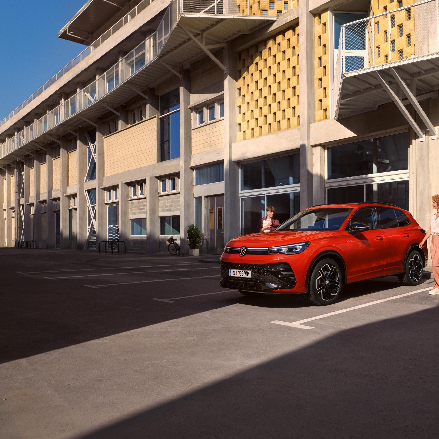 Červený VW Tiguan z bočného pohľadu stojí pred budovou. K vozidlu prichádza žena a chlapec z opačnej strany. Za vozidlom je budova, ktorá vyzerá ako škola.