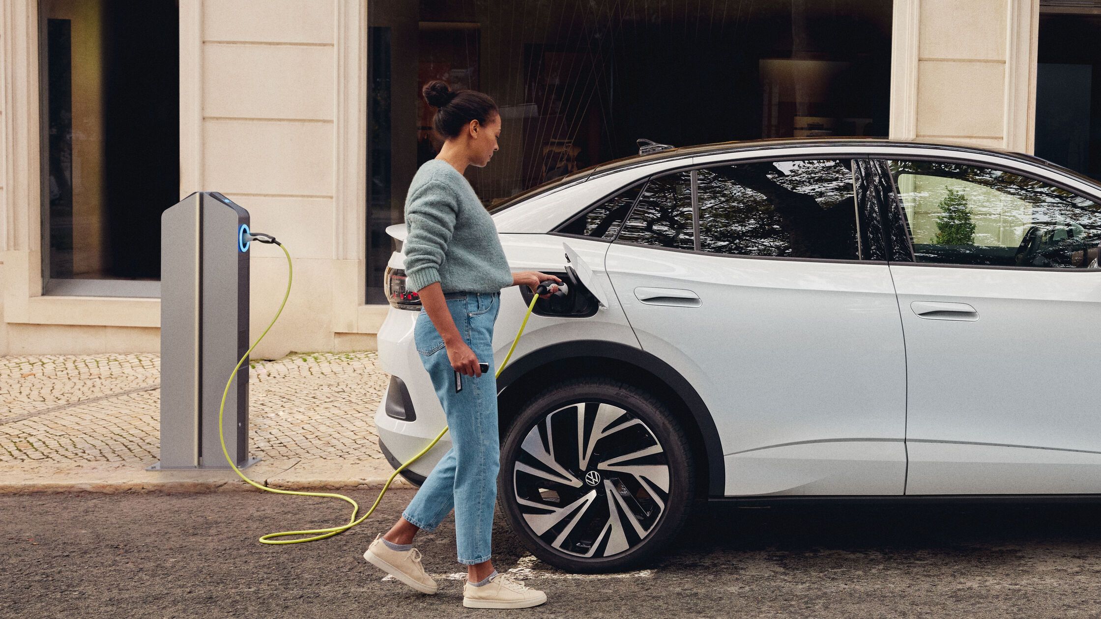 Žena zaparkovala svoj Volkswagen ID. pri nabíjacej stanici a zapojila nabíjací konektor do vozidla. Vozidlo stojí na ceste pri budove a nabíja sa.
