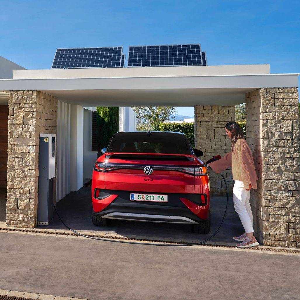 VW ID.5 GTX zaparkovaný pod prístreškom pri dome je napojený na domácu nabíjaciu stanicu a žena pripája k vozidlu nabíjací kábel. 