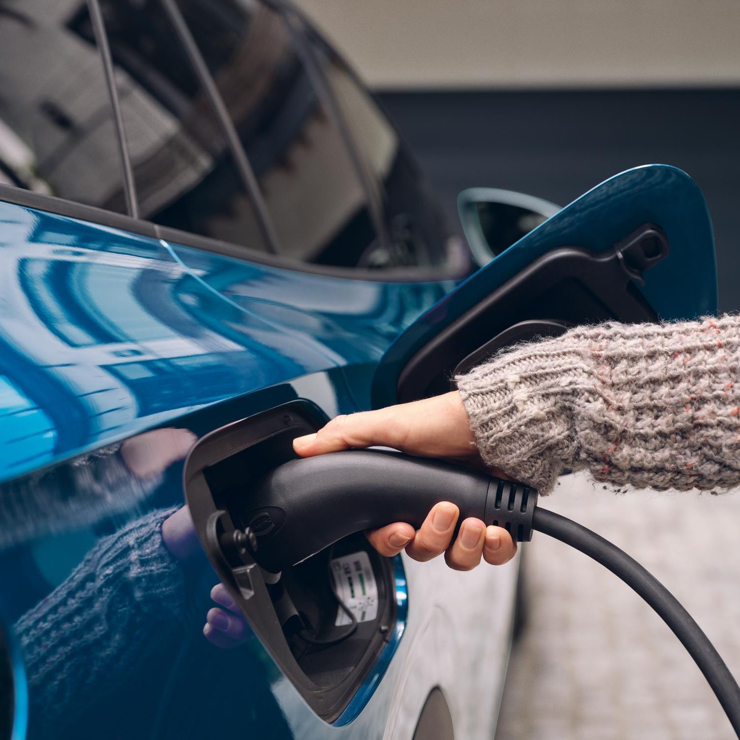 Osoba zapája nabíjací konektor do elektrického auta. Auto je modrej farby.