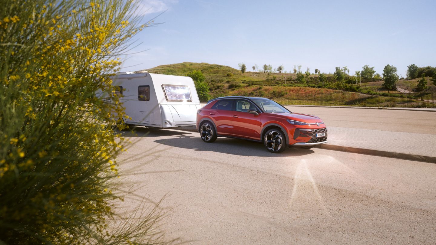 Červený T-Roc ťahá biely karavan po asfaltovej ceste. Krajina je slnečná a v pozadí sú vidieť zelené kopce. Na ľavej strane obrázku je vidieť ker so žltými kvetmi.